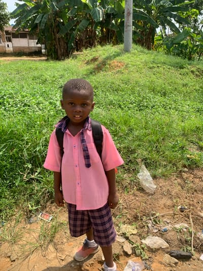 A young boy stands centrally in a full-frame, eye-level outdoor shot in Anantigha, Nigeria. He is of dark complexion, with short, dark, textured hair, and directly faces the camera with a neutral expression, mouth slightly ajar. He wears a light pink short-sleeved collared shirt with a blue and white checkered tie loosely draped around his neck. A black backpack is visible on his shoulders. He wears blue and white checkered shorts, light pink and white sneakers on his left foot, and a white sneaker on his right foot.
The immediate foreground consists of bare dirt and sparse green vegetation, scattered with debris including crumpled plastic bags, wrappers, and small rocks. To the right of the boy, a crushed clear plastic bottle is visible on the ground. Behind the boy, a grassy embankment slopes upwards, covered in various green plants. A grey utility pole with faint markings is positioned on the right side of the embankment. In the background, beyond the embankment, a dense cluster of large-leafed green plants, consistent with banana trees, is present. To the far left background, a light-colored, single-story building with multiple window openings is partially visible amidst the foliage. The scene is illuminated by natural daylight, casting a shadow from the boy's head and upper body downwards and slightly to the right.