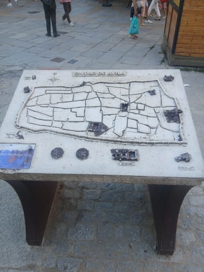 The image shows a relief map of the city of Ávila, Spain, mounted on a stone pedestal in an outdoor public space. The map depicts the city's walls and inner streets, with raised sections representing important buildings or landmarks. Text on the map reads "CIUDAD DE AVILA". To the left of the map, a blue plaque is visible, with partial text including "Smart Heritage City". Two circular medallions, possibly informational markers or decorative elements, are also present on the map's surface. The pedestal is constructed from stone and supported by two dark, curved legs. In the background, paved ground with stone slabs is visible, along with the legs of a few people walking. A wooden structure with a dark roof is located to the right of the scene. The lighting suggests it is daytime.
