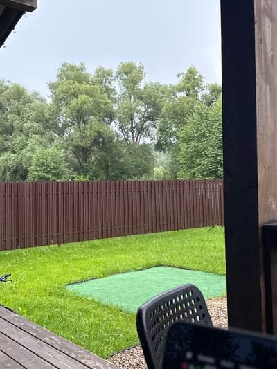 The image shows a backyard scene in Gavshino, Russia, under an overcast sky. In the foreground, a wooden deck partially occupies the bottom left corner, with a black mesh chair and a blurred dark object, possibly a laptop, visible on the right. Beyond the deck, a well-maintained green lawn stretches across the yard. A patch of artificial green turf is situated on the lawn. A brown metal fence runs across the middle ground, separating the lawn from a dense line of lush green trees in the background. A garden hose with a blue nozzle is visible on the grass near the fence. The overall impression is one of a tranquil, domestic outdoor space.