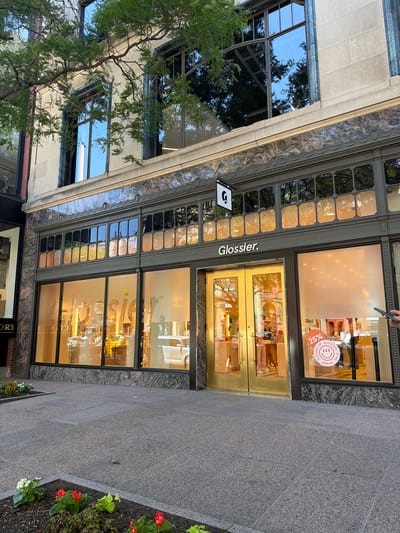 A street-level view captures the exterior of a Glossier store in Boston, United States of America, on a clear day. The store's facade features large glass windows that display the interior, illuminated with a warm, orange glow. The name "Glossier" is prominently displayed above the entrance, which is flanked by golden doors. A sign with a white "G" logo hangs above the storefront. The building's upper floors have large, multi-paned windows reflecting the sky and surrounding trees. Decorative stonework and metallic detailing adorn the building's architecture. The sidewalk in front of the store is paved with grey granite tiles. Planter boxes with red and white flowers are visible along the edge of the sidewalk. In the window to the right, a circular red and white sticker advertises a "25% FRIENDS OF GLOSSIER ANNUAL SALE" with a smiling emoji. A person's hand holding a smartphone is visible on the far right of the image, partially obscuring the window display. The scene depicts a commercial establishment in an urban setting.