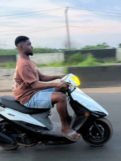 A young Black man is riding a white scooter in Orifite, Nigeria. He has a short, faded haircut and is wearing a brown t-shirt and light blue denim shorts. The scooter has a yellow headlight. The background shows a blurred landscape with greenery and power lines against a cloudy sky, suggesting an outdoor, possibly semi-urban or rural environment. The scooter is in motion on a paved surface, indicated by the motion blur.