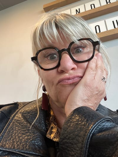 A person with white hair is facing the camera, wearing eyeglasses, a ring, and a black leather jacket. The person's right hand is touching their forehead. In the background, there is a set of white panels with black lettering. The location is unknown, and there is no indication that this image was taken in Andrézieux-Bouthéon, France.