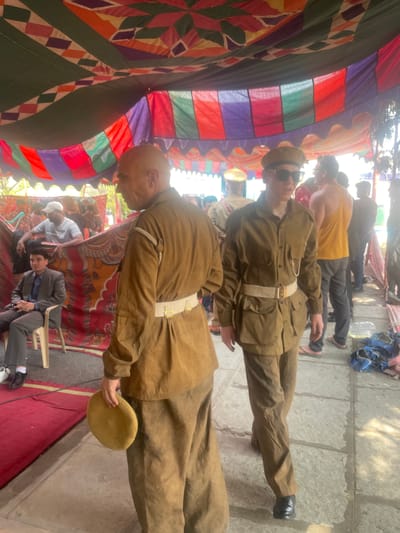 The image captures a scene in Anajpur, India. Underneath a vibrant, patterned tent canopy, two individuals are dressed in tan uniforms with white belts and hats. The foreground figure, bald, holds a tan object in his hand. Behind them, a person in a yellow tank top is visible, along with another in a suit sitting on a chair. The ground is a mix of stone pavement and a red rug. Other people can be seen in the background, some seated and some standing.