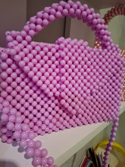 A close-up, eye-level shot features a light purple beaded handbag with a matching handle. The handbag is made of numerous small, round, shiny purple beads that are strung together in a grid-like pattern. The handle is also constructed from the same beads, forming a curved loop. To the right of the primary subject, a portion of another beaded bag, in a slightly darker pink or rose hue, is visible. This second bag also appears to be made of beads and has a curved handle. In the background, a blurry yellow object, possibly a container, and the edges of some colorful items, such as pens or pencils, are partially visible on a shelf. The setting is a shelf, suggesting an indoor environment. There are no people or animals in the frame, and no action is taking place. The lighting is even and appears to be artificial, highlighting the shiny texture of the beads. The specific location within the city of Korenovsk, Russia, cannot be determined from the visual information provided in the image.