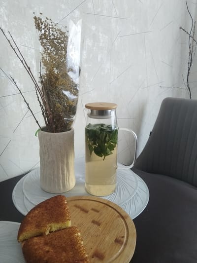 In a dimly lit room, possibly a home interior in Bezlesnyy, Russia, a still life is arranged on a dark table. A clear glass pitcher filled with a pale yellow liquid and fresh green mint leaves sits prominently. Next to it, a cream-colored ceramic vase holds a bouquet of dried branches and flowers, partially encased in clear plastic. In the foreground, two slices of a golden-brown, textured pastry are placed on a circular wooden cutting board. The backdrop features a wall with a textured, white surface adorned with subtle geometric lines. A portion of a dark gray upholstered chair is visible on the right side of the frame, suggesting a seating area. The overall atmosphere is quiet and domestic.