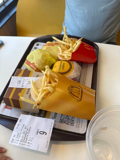 A close-up, overhead shot shows a fast-food meal on a black plastic tray. The meal includes two burgers, a red box of French fries, and a yellow fry holder, all with McDonald's branding. A receipt is visible at the bottom of the tray, listing items ordered: 1 L Coke, 1 M Fanta Orange, 2 Bacon Dbl QPC, 2 Cheeseburger, 1 Double Cheese, 1 M Fry, and 1 L Fry. The table number is 9. A clear plastic cup with some liquid is partially visible on the right side of the tray. The background shows a yellow wall and a person wearing a light blue t-shirt. The scene appears to be inside a McDonald's restaurant, possibly in Placerville, United States of America, given the context provided.