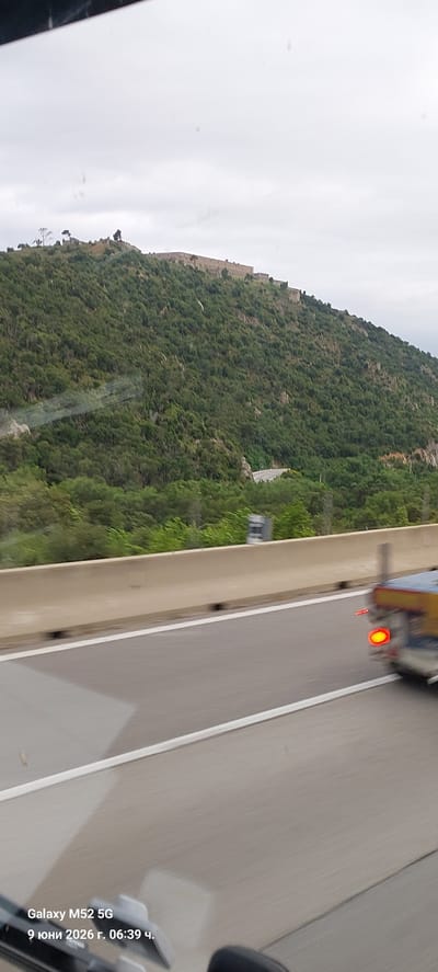 The image is a photograph taken from inside a vehicle, showing a landscape view. In the background, a large, stone fortification or castle is visible on top of a heavily wooded hill. The hill is covered in dense green trees. Below the hill, a winding road is visible, and in the foreground, a highway barrier separates the road from the camera's viewpoint. Part of a vehicle with a lit red taillight is visible on the highway. Text overlay on the bottom left corner indicates the device used ("Galaxy M52 5G") and a date and time ("9 June 2026, 06:39"). The scene is outdoors, under an overcast sky. This landscape is consistent with the area around La Jonquera, Spain, a town known for its location near the French border and surrounding natural terrain with historical fortifications.