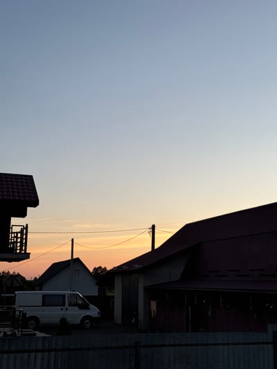 This is a photograph of a rural scene in Chemerne, Ukraine, taken during sunset. The image is oriented vertically. The sky takes up the left two-thirds of the frame, transitioning from a pale blue at the top to a soft orange and yellow gradient closer to the horizon, indicating the sun is setting or has recently set. A few wispy clouds are visible in the sky.
The right third of the image features several structures. A large, dark-colored building with a corrugated roof dominates the upper right. Below it, there are smaller residential buildings, one with a dark, sloped roof. A white van is parked in front of one of these buildings. A utility pole with wires is also visible, stretching across the frame horizontally. The scene suggests a quiet, possibly residential or industrial area on the outskirts of a town. The time of day is clearly evening, characterized by the warm sunset colors in the sky. The weather appears clear, with no rain or strong winds suggested. No people are visible in the frame, and there is no visible text.