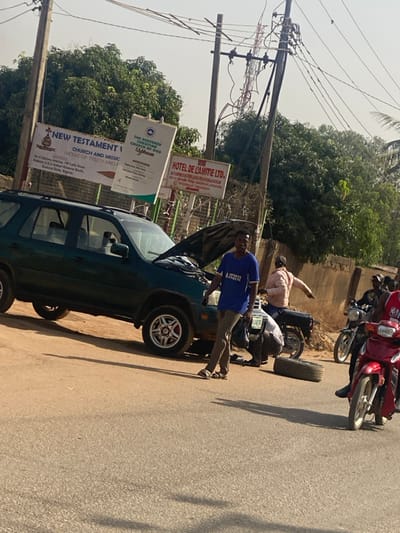 The image depicts a roadside scene in Makera, Kaduna State, Nigeria. A dark green Honda CR-V SUV is parked partially off the paved road on the left, with its hood open and the front left wheel removed. The removed tire rests on the ground beside the vehicle. A male individual, wearing a light-colored shirt, is crouched near the front left wheel area, appearing to work on the SUV. Another male, dressed in a blue t-shirt with "INFINITE ATHLETE" text, brown trousers, and sandals, stands upright near the SUV's front. On the paved road to the right, two motorcycles are present; one dark, ridden by a male in a light-colored shirt, and one red, ridden by a male in a red shirt.

In the background, three prominent signboards are visible: "NEW TESTAMENT CHURCH AND MISSION (HOUSE OF TRUTH AND FAITH)," "THE REDEEMED CHRISTIAN CHURCH OF GOD," and "HOTEL DE L'AMITIE LTD.", all displaying addresses referencing Makera. Overhead utility poles with numerous wires are present, and a tall cellular tower is visible in the distance. The environment is characterized by a paved road, a dirt shoulder, green foliage from trees, and a clear, bright sky.
