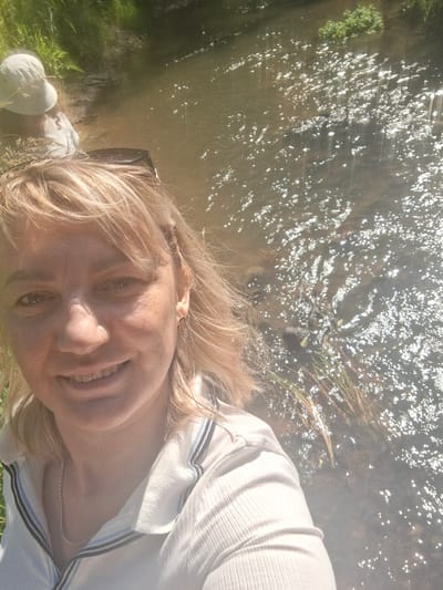 A woman with blonde hair and fair skin takes a selfie by a body of water. She is smiling and wearing a white collared shirt with blue and black stripes on the collar. Her hair is styled with a braid on the right side. Behind her, another person wearing a white hat is partially visible near the edge of the water. The water is shallow and reflects sunlight, creating a shimmering effect. There is vegetation along the bank of the water. The scene appears to be a natural setting, possibly a stream or shallow river.