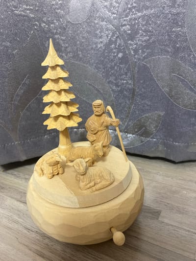 The image shows a carved wooden nativity scene. The scene is set on a circular wooden base. On the base, there are three carved sheep, a carved shepherd holding a staff, and a carved Christmas tree. The background is a grey textured fabric with a floral pattern. The scene is placed on a wooden surface. The image is taken from a slightly elevated angle.