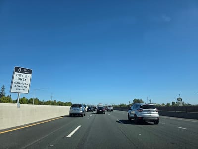 A view from inside a vehicle traveling on a highway. The sky is clear and blue. A concrete barrier runs along the left side of the road, and a diamond-shaped sign indicates "LEFT LANE HOV 2+ ONLY" with specific hours of operation. Several cars are traveling in the same direction on the highway, including a silver SUV in the foreground, a dark sedan, and a white truck. Trees and power lines are visible in the background, along with a distant sign structure. The road is asphalt with white lane markings. The license plate of the silver SUV is visible and reads "8TGW694". The scene appears to be in Elk Grove, California, United States of America, based on the context of the HOV lane sign common in that region.