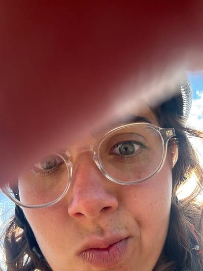 The image displays a close-up, upward-angled selfie of an individual's face, predominantly a female, partially obscured by a reddish-brown object, likely a finger, in the upper left quadrant. The individual has fair skin, green eyes visible through clear, round-framed spectacles, and dark hair, part of which is visible on the right side. A dark, possibly ribbed object resembling a helmet is partially visible on the right side of the head. The individual's lips are slightly pursed, forming a pout. Strong sunlight illuminates the right side of the face and hair, indicating an outdoor setting. A patch of blue sky with white clouds is visible in the upper right corner. No discernible features or landmarks within the image identify the location as the city of Nadao, Laos.