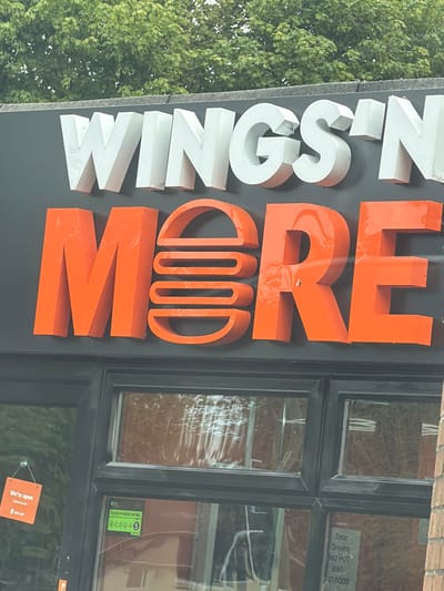 The image depicts a storefront in Pity Me, United Kingdom. A sign reading "WINGSIN MORE" is prominently displayed, with "WINGSIN" in white letters and "MORE" in orange. A window next to the sign has stickers and signs, including a food hygiene rating of 5, a note to delivery drivers, and an "open" sign advertising Just Eat. Foliage of a tree is visible in the background. The location appears to be outdoors.