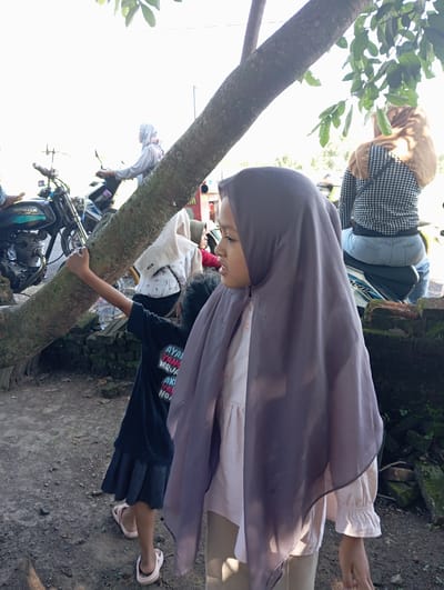 A vertical, ground-level shot shows a young girl in a dark blue dress and light pink sandals, gripping a tree trunk. The girl, who is wearing a hijab, looks down. A dirt path surrounds the tree. Behind her is a motor bike with an adult seated on it. A second motor bike is parked to the left of the tree. Several people are in the background. The location is in Waung, Indonesia.
