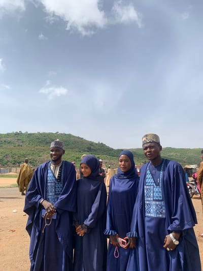 Four individuals are captured in an outdoor photograph in Naraguta Hausawa, Nigeria, taken during daylight under a partly cloudy sky. Two men, positioned at the top and bottom of the frame, and two women, located in the middle, are all wearing similar, flowing blue garments. The men have matching hats, while the women have head coverings. The men are holding mobile phones and prayer beads, and one woman holds a pink smartphone. The background features a green, hilly landscape and a dirt road. Other people and vehicles are blurred in the background.