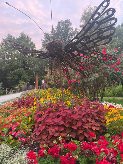 This is a photograph taken in the city of Innsbruck, Austria. The foreground is dominated by a vibrant flower bed featuring a mix of red, pink, yellow, and white blossoms, along with deep red foliage plants. Behind the flowers, a large, intricate metal sculpture of a butterfly is prominently displayed, its wings made of interconnected circular elements. The sculpture is positioned amidst more greenery, including red roses and other bushes. In the mid-ground, a wooden bridge crosses a path, and two individuals are visible on the bridge. One person, wearing a dark jacket and jeans, stands looking towards the left. The other person, wearing a lighter colored shirt, stands closer to the right side of the bridge, holding what appears to be a mobile phone to their ear. Trees and a cloudy, pastel-colored sky form the background, suggesting it may be dusk or dawn. A lamppost is visible near the bridge. The scene appears to be a park or garden setting.