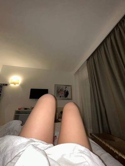 A person lies in a bed, covered with a white blanket. Their bare thighs are visible. The bed is situated in a hotel room. A picture hangs on the wall above a flat-screen television. A small lamp provides light near the bottom of the frame. Neutral-colored curtains can be seen at the top. The perspective is from a low angle, looking up and slightly toward the right.
