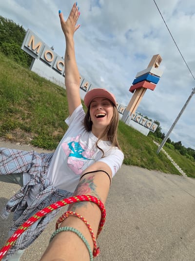 A young woman is taking a selfie on a cloudy day in Nikolaevka, Russia, in front of a large sign that says "MOSCOW". She is wearing a pink baseball cap, a white t-shirt with a graphic design, and a plaid shirt tied around her waist. Her right arm is raised in the air, and she is smiling broadly. She has tattoos on her arm and is wearing bracelets. The sign is large and white with orange letters. Behind the sign, there are trees and a grassy hill. There is also a tall pole with colored flags on it. The ground in front of the sign is asphalt.