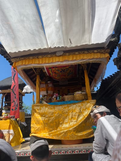 This is an eye-level, medium shot of an outdoor religious ceremony in Mendoyo, Indonesia. The scene is dominated by a decorated altar, likely for a Balinese Hindu ceremony. The altar is draped in vibrant yellow fabric with intricate gold floral patterns. Above the altar, white fabric hangs like a canopy, billowing slightly in the breeze, with a clear blue sky visible through it. The altar itself holds various offerings, including woven baskets, what appears to be food items, and possibly incense. To the left of the altar, a tall red banner with a dark design is visible, and further back, a dark, dome-like structure. In the foreground, three men are partially visible, facing the altar. One man in the immediate foreground wears a white head covering and a white long-sleeved shirt. His gaze is directed towards the altar. The overall atmosphere suggests a day of prayer or celebration.