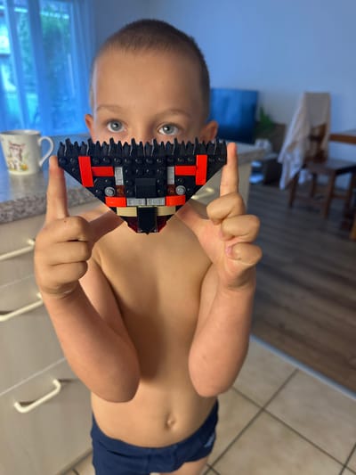 A young boy is holding a black and red LEGO creation up to his face. The LEGO structure is triangular in shape with some silver and tan colored pieces incorporated. The boy's eyes are wide open and looking directly at the camera. He appears to be in a home environment, with kitchen cabinetry visible in the background. The lighting suggests it could be daytime. The image is oriented vertically.