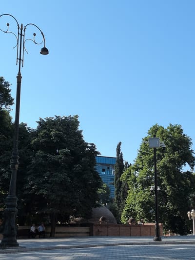 The image depicts a street scene in Ganja City, Azerbaijan, likely in a park or public square. A decorative, ornate lamppost stands prominently on the left side of the frame. In the background, dense green trees provide shade, and a modern building with a glass facade is visible. To the right, another lamppost, this one equipped with security cameras, is situated amongst the trees. In the middle ground, two individuals are seated on a bench under the shade of a tree, facing away from the camera. A low brick wall with dome-like structures is also visible. The ground in the foreground appears to be paved with light-colored stones, suggesting a pedestrian area. The sky is clear and blue, indicating a sunny day.