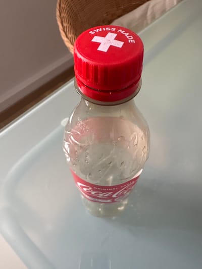 The scene is located in the city of Visp, Switzerland. A clear plastic Coca-Cola bottle stands upright on a light-colored, reflective surface. The bottle is approximately one-quarter full of a clear liquid and exhibits condensation droplets on its interior. A red label segment with the "Coca-Cola Original Taste" branding is visible on the bottle's lower half. The bottle is capped with a red, ribbed screw-top lid. The top surface of this cap features a white Swiss cross centrally positioned, surrounded by the white text "SWISS MADE" in an arc. In the blurred background, a woven brown material, consistent with a wicker basket, is partially visible, along with indistinct lighter surfaces. No individuals or active interactions are depicted.