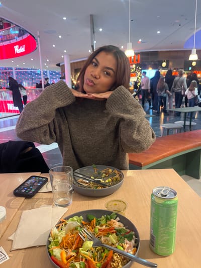 An indoor photograph depicts a female subject seated at a light wooden table within the Westfield Mall of Scandinavia, Solna, Sweden. The subject, positioned centrally, has medium skin tone and dark brown hair with reddish highlights. She wears a dark grey knitted sweater and rests her hands under her chin, making a playful facial expression with her tongue slightly extended. A silver ring is visible on her left ring finger.

On the table in the foreground, from left to right, are a black smartphone displaying app icons, a clear glass filled with water, a folded light brown napkin, two grey bowls containing partially consumed salads, a small clear plastic cup with sauce, and an open light green aluminum beverage can. One salad bowl is nearly empty, while the other contains a mix of lettuce, shredded carrots, red bell peppers, edamame, and other ingredients, with a fork resting within.

The background shows a brightly lit mall environment. To the left, a large red LED screen displaying the "Westfield" logo is visible above an ice rink where several indistinct figures are engaged in skating. To the right, numerous blurred individuals are seen walking and standing, indicative of a busy public space. Overhead lighting fixtures and architectural elements such as pillars are present.