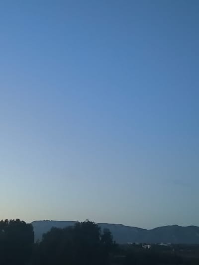 The image displays a panoramic twilight landscape, likely captured from Nicosia, looking north towards the Kyrenia Mountains (Pentadaktylos range). The upper approximately two-thirds of the frame consists of a clear, gradient blue sky, transitioning from a lighter hue at the horizon to a deeper blue overhead. The lower third of the image shows a distant, dark silhouette of a mountain range. Prominently situated on the ridge of the leftmost visible mountain, a series of bright, illuminated white lights are discernible, forming what appears to be a large emblem or structure. In the foreground, below the mountain range and extending across the lower portion of the image, are dark, amorphous silhouettes of trees and vegetation. Faint, scattered artificial lights indicative of urban or suburban development are visible in the mid-ground, particularly towards the right side, nestled within the valley or lower slopes. No individuals, specific actions, or close-up objects are present in the frame.