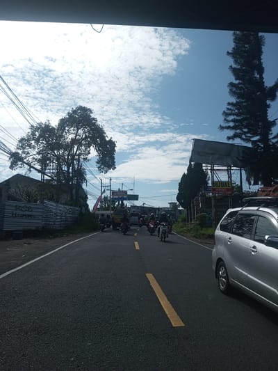 The image depicts a street scene in what appears to be a Southeast Asian location, possibly Khe Luang, Laos. A grey asphalt road runs through the center of the frame, marked with a broken yellow line. Several motorcycles are visible on the road, some moving towards and others away from the camera. On the right side of the road, a silver car is partially visible. A large tree stands on the left side of the road, with utility poles and wires obscuring some of the view. A building with a metal fence and possible graffiti is on the left. On the right, there are tall trees, signs, and buildings. The sky is partly cloudy with blue patches.