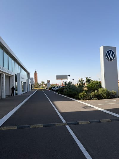 The image shows an outdoor view in Ouahat Sidi Brahim, Morocco, oriented vertically. A Volkswagen dealership sign is prominently displayed. A modern building with glass windows features signs in French. A road marked with white lines leads away from the building. Other commercial buildings, including a Porsche dealership sign and a tower, are visible in the background. A person is standing near the entrance of the building. Vegetation lines the side of the road. The sky is clear and blue.