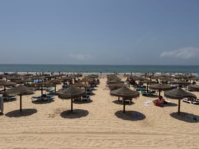 This is an eye-level, full shot of a crowded beach in Vila Real de Santo António, Portugal. The foreground is dominated by rows of thatched umbrellas and empty lounge chairs on a sandy beach. In the mid-ground, more umbrellas and chairs are occupied by beachgoers, some of whom are sunbathing or sitting. The background features the Atlantic Ocean, with many people swimming and wading in the water. The sky is a clear, pale blue, with a few wispy clouds on the horizon. The overall impression is of a vibrant and busy beach on a sunny day.