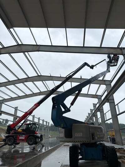 The image depicts an active construction site in Stróża, Poland. Two aerial work platforms are positioned beneath the skeletal framework of a large building under construction. A red Manitou telescopic handler is parked on the wet concrete floor in the foreground, its stabilizers extended. Adjacent to it, a blue Genie Z-30/60 boom lift is raised, with its basket positioned near the steel beams. A person is visible in the basket of the boom lift, appearing to be working on the steel structure. The background reveals more of the unfinished building's steel frame, an overcast sky, and a wooded area with some construction materials scattered around. The ground is wet and reflective, suggesting recent rainfall.