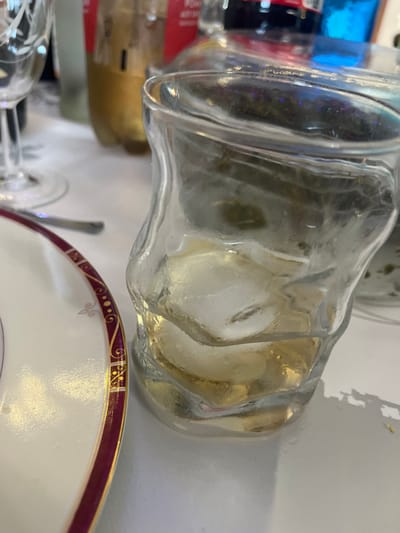 The scene is set on a white tabletop in Ancenis, France. A clear, irregularly shaped glass tumbler occupies the central foreground, containing a pale yellow liquid and approximately three discernible ice cubes. To the immediate left, a portion of a white ceramic plate is visible, featuring a decorative dark red rim adorned with gold scrollwork and dots. A silver-toned utensil handle is positioned between the plate and the tumbler. In the blurred background, several bottles are present: one appears amber-colored, another exhibits a red label, and a third shows a distinct blue hue. A stemmed, clear glass with etched patterns is partially visible on the far left. No individuals or direct actions are depicted.