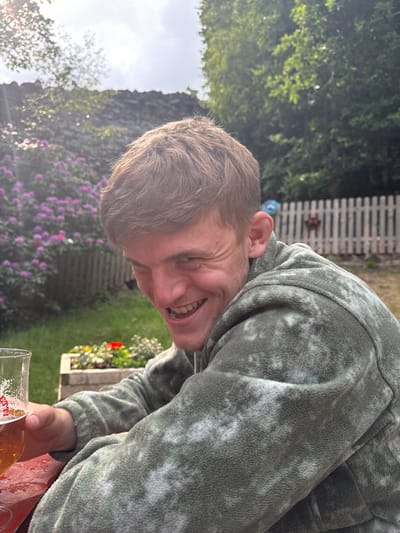 A young, caucasian male, wearing a green, white, and gray tie-dye fleece jacket, is holding a glass of amber liquid in his right hand. He is looking at the camera and smiling. The man is sitting at a wooden table outdoors, in a garden setting. There is a wooden fence behind him, and a patch of pink and purple flowers to his left. The man is in the foreground, and the background is slightly out of focus. The scene is located in Hayfield, United Kingdom, at the coordinates 53.377 latitude and -1.935 longitude.