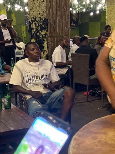 The image depicts a group of people gathered in what appears to be an outdoor seating area with lush green foliage and decorative lighting overhead, suggesting a restaurant or lounge in Oba, Nigeria. In the foreground, a man is seated, wearing a white t-shirt with text and a gold chain. He is holding a black object in his lap. To his left, on a wooden table, are two green glass bottles, one appearing to be a Heineken. In the background, other individuals are visible, some seated and engaged in conversation. The overall ambiance is casual and social.