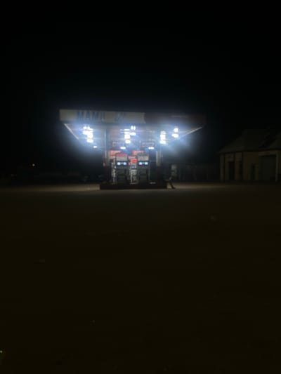 At night in Unguwar Fatika, Nigeria, a brightly lit “Mami Oy” convenience store/gas station is visible on the left side of the frame. The right side of the frame is dark.