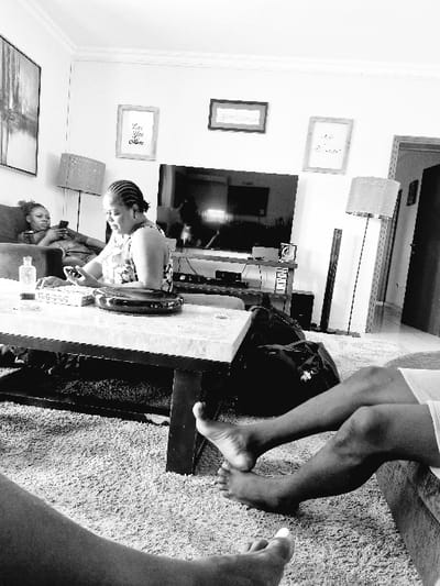 An interior scene captured in black and white presents a family at home. A woman with braided hair leans over a table, focused on an object in front of her. To the side, a young girl reclines on a sofa, possibly engaged with a handheld device. Legs are visible, suggesting someone is nearby, perhaps seated on a high surface. Framed artwork hangs on the wall, adding a personal touch to the space. The carpet adds texture and depth.