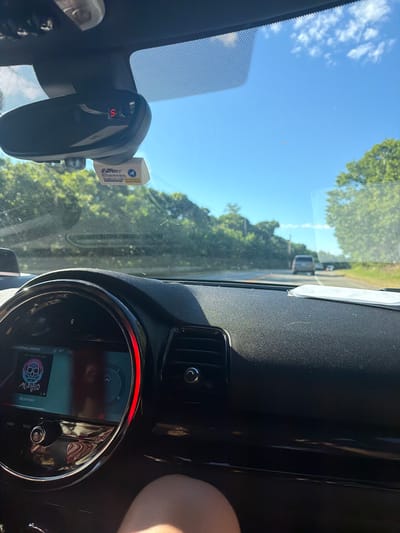 The image is a first-person view from inside a car, looking forward through the windshield. The dashboard is visible in the foreground, featuring a circular digital display with a red illuminated outer ring, showing "BLUETOOTH: SPOTIFY" and album art of a skull with headphones. A small digital display above the rearview mirror shows the number "5" in red. An EZ-Pass transponder is attached to the windshield. The car is on a paved road, with trees and greenery lining both sides. Other vehicles are visible on the road ahead, including a dark SUV. The sky is bright blue with scattered clouds. The image was captured in Truro, Massachusetts, United States of America.