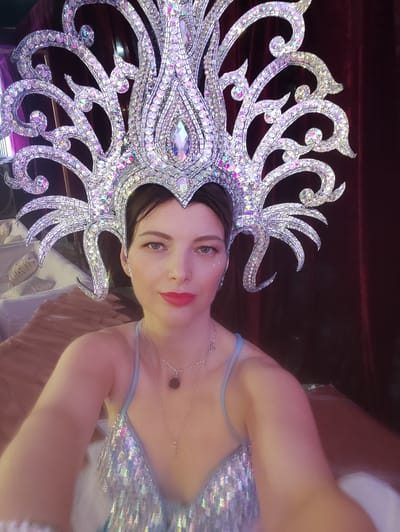 A woman wearing an elaborate, shimmering costume poses for a close-up, likely a selfie or portrait, in an indoor setting, with the provided context suggesting Askihala, India. She looks directly at the camera with a calm, composed expression.
Her most striking feature is a large, ornate headdress made of silver-colored material, intricately adorned with countless iridescent beads and larger crystals that reflect light in various hues of purple, pink, and blue. The headdress features a prominent central pointed element and multiple swirling, decorative extensions that frame her face and extend upwards and outwards.
She has dark hair pulled back, visible beneath the headdress. Her makeup includes bold red lipstick and subtle glitter or small jewels applied to her left cheekbone. She is wearing a light blue, thin-strapped top that is heavily embellished with matching iridescent sequins, complementing the headdress. Two delicate silver necklaces with small pendants, one dark and circular, the other star-shaped, are visible around her neck.
The background is softly lit and blurred. To her right and behind her is a rich, dark red velvet curtain, suggesting a theatrical or performance venue. To her left, part of a lighter, indistinct object or piece of furniture is visible. The overall lighting is dim but effectively highlights the reflective qualities of her dazzling costume, implying she is dressed for a performance or special event. No visible text is present in the image.
