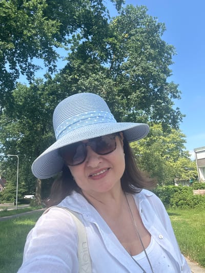 A woman is taking a selfie outdoors. She is wearing a blue straw hat with a white polka dot ribbon, sunglasses, and a white linen shirt over a white tank top. A silver necklace is visible. She is standing in front of large green trees and a bright blue sky. In the background, there is a street with a lamppost, houses, and green grass. The image appears to be taken in a suburban or park-like setting in Steenwijk, Netherlands.