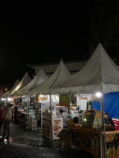 A nighttime view shows a street market lined with vendor stalls under white peaked canopies. People browse and work at the stalls, which are stocked with various goods and food items. Signage is visible, advertising "Sari Jeruk Manis" (sweet orange juice) and other products. The scene is illuminated by overhead lights, and the ground is paved with bricks. The overall atmosphere suggests a vibrant and busy outdoor market.