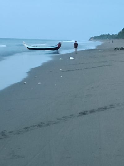 The image depicts a tranquil scene on a beach in Curehtunong, Indonesia, during what appears to be either dawn or dusk, as indicated by the soft, diffused light. A traditional Indonesian fishing boat, with its distinctive curved hull and outriggers, rests partially on the wet sand at the water's edge. The boat is dark in color with a lighter-colored hull. A figure, wearing a red garment and dark pants, is positioned near the stern of the boat, suggesting they may be tending to it or preparing for a voyage.
Further down the beach, a single individual, dressed in a dark t-shirt and light-colored pants, is walking away from the water and towards the interior of the beach. Their posture indicates a casual stroll. The beach itself is composed of fine, light-colored sand. In the distance, a line of lush green trees forms a dense backdrop, with a hint of distant lights suggesting a settlement or activity beyond the immediate coastline. The ocean is calm, with gentle waves lapping at the shore. The overall atmosphere is serene and peaceful, conveying a sense of everyday life in this coastal Indonesian community.