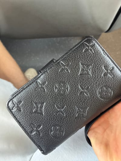 A person is holding a black embossed wallet with a repeating pattern. The wallet has a textured surface and a slightly shiny finish. The background is blurred and appears to be a seat or bench, suggesting the photo was taken indoors. The person's hand is visible holding the wallet. There is no indication of the location being Móstoles, Spain.