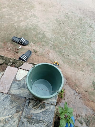 The image depicts an outdoor scene in Gindin Dutse, Nigeria. The primary subjects are two pairs of flip-flops and a green plastic bucket filled with water. One pair of black flip-flops with white stripes is situated on a dirt and concrete ground, while another similar pair lies further down on the same surface. A third, smaller flip-flop, yellow with blue accents, is partially visible near the bucket.
The green bucket, containing a noticeable amount of water, sits on a patterned tiled surface, which transitions to dirt. The ground is a mix of wet dirt and dry, dusty patches, suggesting recent rain or a water spill. Water is seen pooling and running across the tiled area. A small, green plant is growing near the bucket, adding a touch of nature to the scene.
The overall impression is one of a utilitarian outdoor area, possibly a yard or a pathway. The lighting suggests it could be daytime, but the overcast conditions might obscure specific time-of-day cues. The presence of water and the wet ground indicate recent precipitation or a source of water, perhaps for domestic use or gardening. There is no visible text in the image. The scene is devoid of any people, and the arrangement of objects suggests a moment of pause, with items left out in the open.