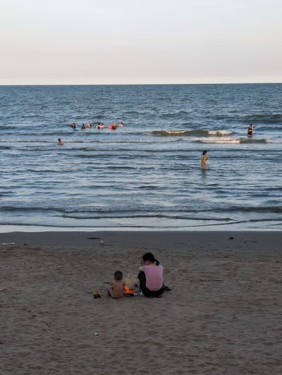This image depicts a beach scene in Hoằng Tiến Commune, Vietnam. In the foreground, a woman in a pink shirt and dark pants sits on the sand with her back to the camera, facing a young child who is also sitting on the sand. The child is facing away from the camera and appears to be playing with toy vehicles. A woven bag is positioned near the woman.

The middle ground features the sea with gentle waves. Several people are visible in the water. Some are wading, while others appear to be swimming or floating with the assistance of flotation devices. One person is standing in the water further out, wearing a light-colored garment.

The background is dominated by the ocean and a pale sky. The horizon is clearly defined between the water and the sky. The beach itself is sandy and appears to have some scattered debris. The overall lighting suggests it is either early morning or late afternoon.