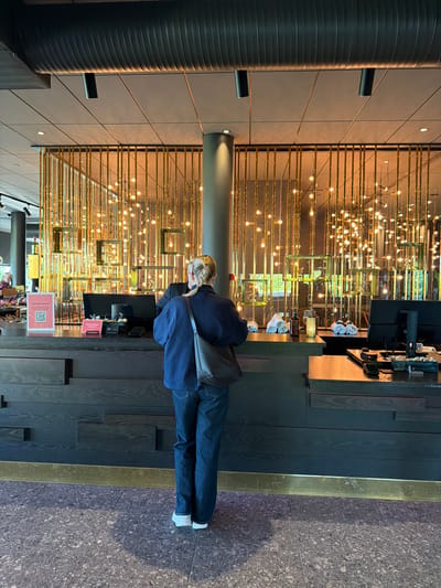 A blonde-haired woman wearing a dark blue jacket and jeans stands at a dark wood counter in what appears to be a reception area. She is facing away from the camera, looking at a computer monitor on the counter. To her left, on the counter, is a pink sign with a QR code and text in Swedish. Behind the counter, a decorative wall is illuminated by numerous golden lights. The floor is a speckled grey tile. The overall impression is that of a modern hotel or commercial establishment. This image was taken in Östersund, Sweden.