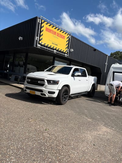 The image shows a white Ram pickup truck parked in front of a dark-colored building with a large yellow sign. The sign has black diagonal stripes and reads "MONTEUR GEZOCHT Fulltime" which translates to "MECHANIC WANTED Fulltime". Below this, it says "Wordt een expert in caravans" (Become an expert in caravans) and provides a phone number.

A person, wearing a white t-shirt and grey shorts, is visible behind the truck, appearing to be working on something attached to another vehicle, possibly a caravan or trailer. The scene is set outdoors on a paved area, likely a parking lot or forecourt. The sky is blue with scattered white clouds, indicating a daytime scene under fair weather conditions. The truck has a Dutch license plate, "V-25-NTF", confirming the location in the Netherlands. The building suggests a commercial establishment, likely a dealership or repair shop for vehicles and caravans. The overall impression is one of a business advertising for staff on a bright, sunny day.
