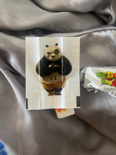 The image shows a sticker of Po from Kung Fu Panda on a wrinkled white sheet, likely a sticker album page, lying on a rumpled grey fabric surface. To the right of the sticker is a partially opened candy wrapper with a yellow emoji winking on it, also on the grey fabric. The fabric appears to be a bedspread or similar textile. There is no visible indication of the location being Hazorasp, Uzbekistan. The image is a close-up of these objects and does not provide any contextual information about its surroundings or location.