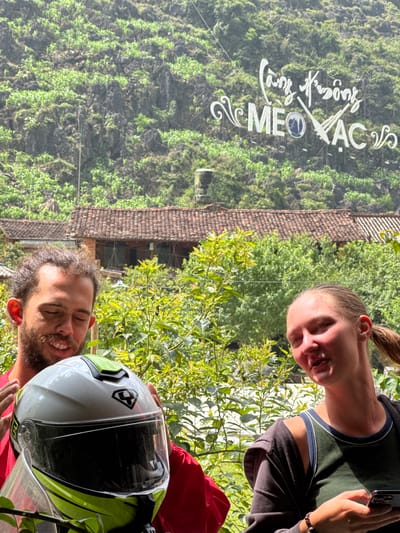 Two people pose for a photo in Mèo Vạc Commune, Vietnam. The woman, with a ponytail and olive shirt, holds a phone. The man with a beard is holding a motorcycle helmet. Behind them is a lush green mountain, the roof of a building, and various vegetation. The location sign "Cung Đường Mèo Vạc" can be seen on the mountain.